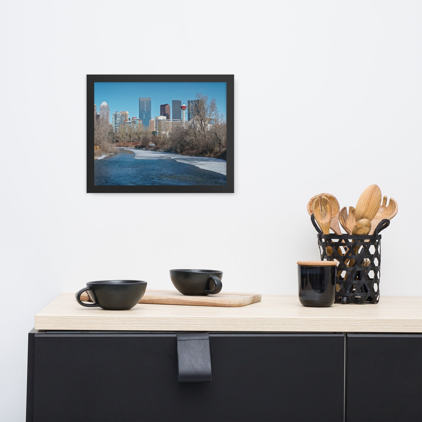 Framed cityscape print above a black and light wood cabinet with cups and kitchen utensils.