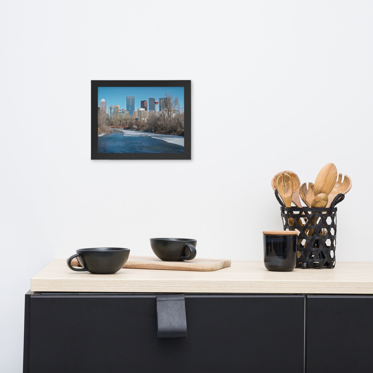 Framed cityscape art above countertop with black cups and utensils.
