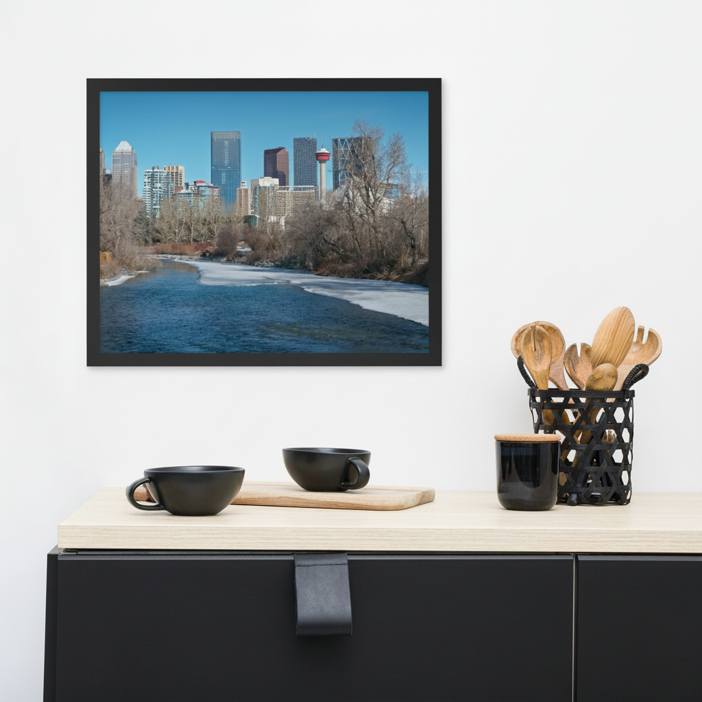 Framed photograph of a cityscape over a river hangs above a kitchen countertop. Black coffee cups, a wooden cutting board, utensils, and a container sit on the countertop. A cabinet door with a leather pull is visible.